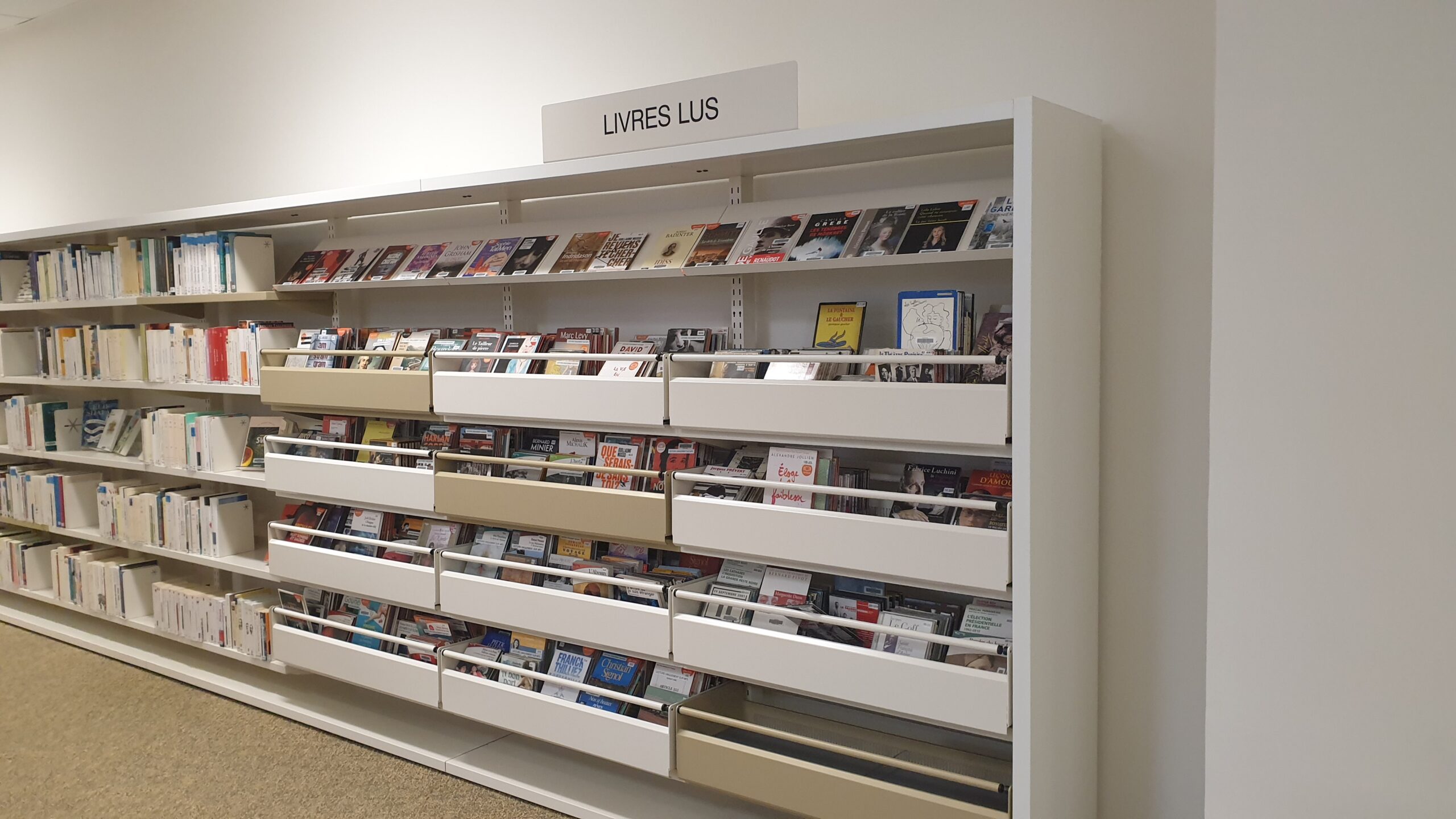 L’image montre un grand meuble de bibliothèque étiqueté « Livres lus », rempli de nombreux livres et CD disposés dans des étagères et bacs inclinés.