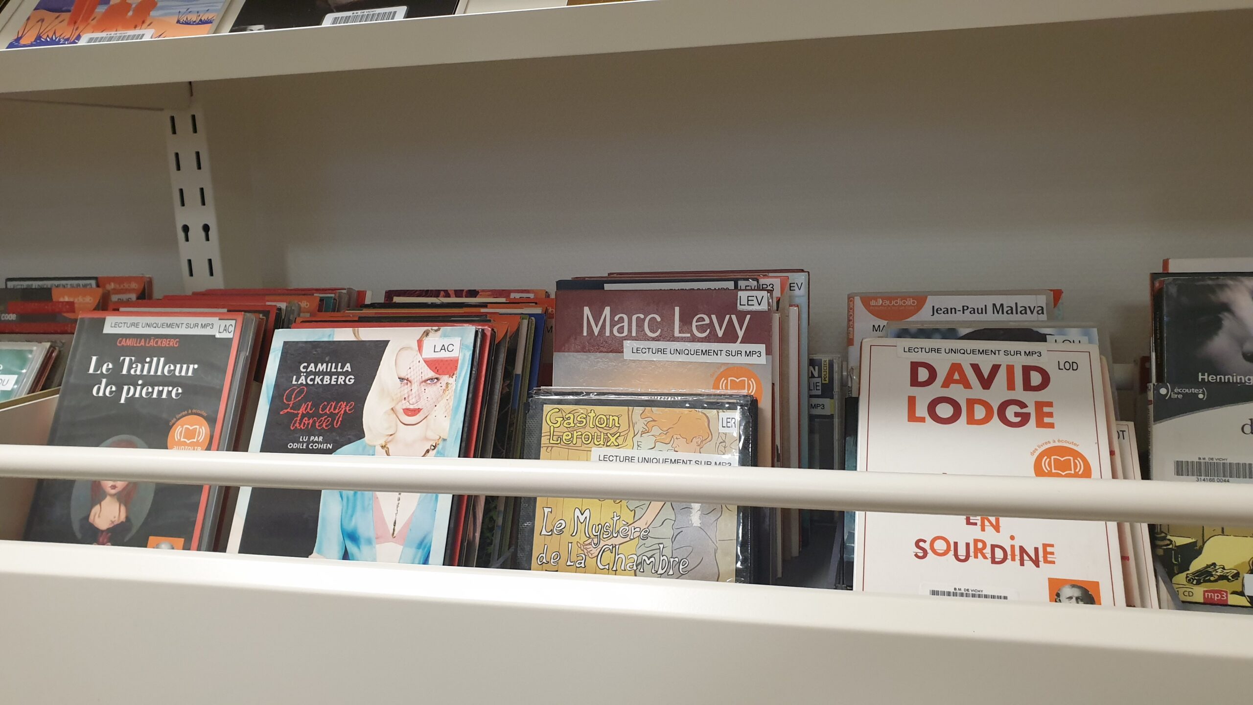 une étagère de bibliothèque remplie de livres audio et documents, avec en avant‑plan plusieurs titres bien visibles rangés dans des pochettes plastifiées.